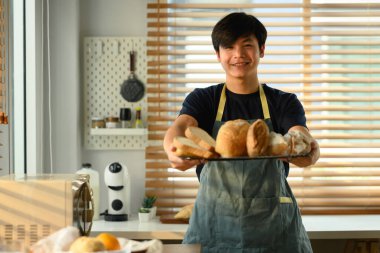 Yakışıklı Asyalı fırıncının portresi. Elinde bir tepsi dolusu taze pişmiş ve kameraya gülümsüyor..
