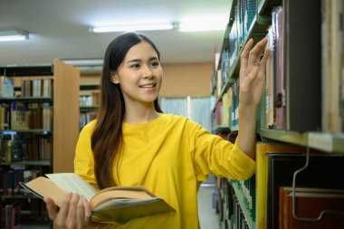 Üniversite kütüphanesinde okumak ve sınava hazırlanmak için raftan kitap seçen pozitif bir üniversite öğrencisi. Eğitim kavramı.