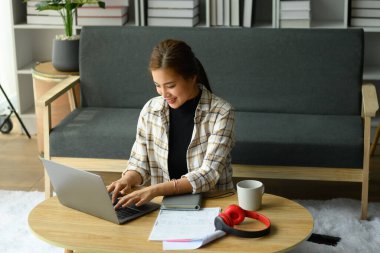 Çekici genç bir kadın, rahat oturma odasında yerde oturuyor ve dizüstü bilgisayar kullanıyor. İnsanlar, teknoloji ve yaşam tarzı.