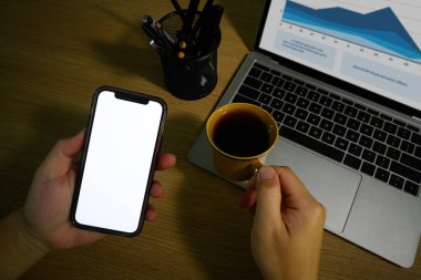 Tanımlanamayan bir elin akıllı telefon ve kahve fincanı tutuş görüntüsü. Reklam metniniz için boş ekran.