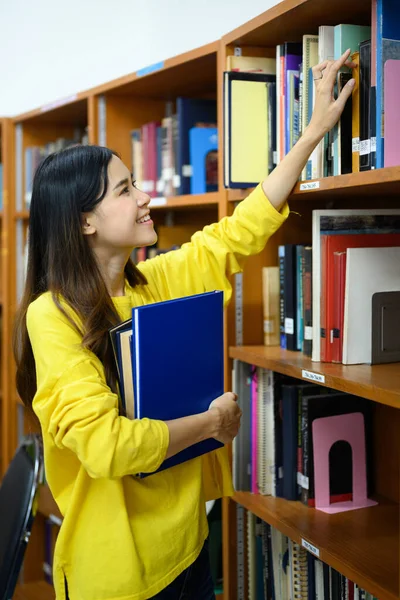図書館で勉強するために棚から教科書を取っている陽気な学生の女性 若者のライフスタイルの概念 — ストック写真