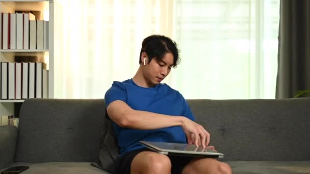 ソファに腰を下ろしてノートパソコンを使うカジュアルな服装のアジア系男性 コミュニケーション テクノロジーの概念 — ストック動画