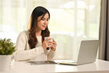 Milenyum Asyalı kadın kahve içiyor, e-posta okuyor, evde dizüstü bilgisayarda çalışıyor..