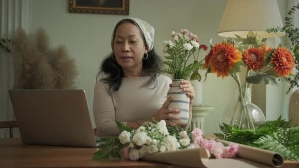 幸せな成熟した女性花屋 小さなビジネスフラワーショップのオーナーは 新鮮な花に囲まれた作業机でラップトップでビデオ通話を持っています — ストック動画