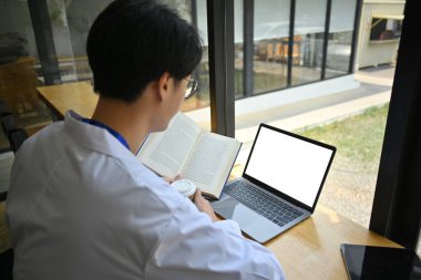 Beyaz önlüklü doktorun elinde kitap tutarken ve hastane kantinindeki ahşap tezgahta dizüstü bilgisayar kullanırken yan görüntüsü.