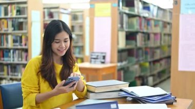 Sosyal medyada sohbet eden hoş bayan üniversite öğrencisi kütüphanede otururken akıllı telefondan eğitim için uygulamalar kullanıyor..