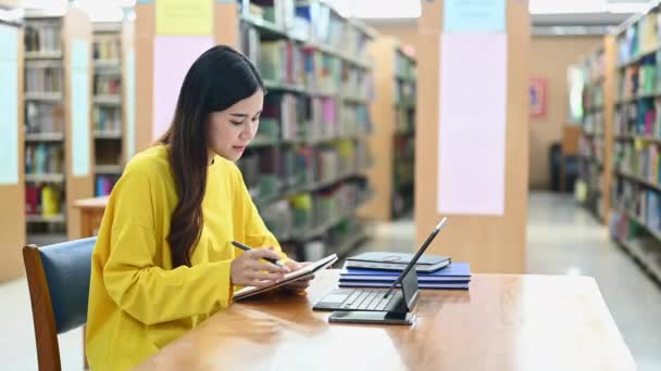 大学図書館に座って宿題をしているアジア系女子学生の笑顔 ノートパソコンで情報を探す — ストック動画