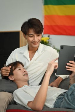 Sevgi dolu homoseksüel çift kanepeye sarılıp dijital tablet kullanıyor. LGBT, aşk ve yaşam tarzı ilişki kavramı.