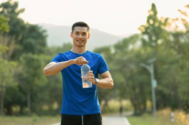 Sportif Asyalı adam elinde bir şişe su tutuyor ve dışarıda egzersiz yaparken kameraya gülümsüyor. Sağlıklı yaşam tarzı, sağlık konsepti.