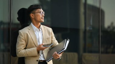 Düşünceli bin yıllık iş adamı akıllı telefon kullanıyor ve iş merkezinin önünde dikilirken gözlerini kaçırıyor..