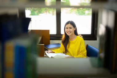 Üniversite kütüphanesinde kitap okuyan neşeli kız öğrenci kitaplığına bak. Eğitim, öğrenme ve gençlik yaşam tarzı kavramı.