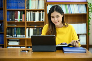 Gülümseyen üniversite öğrencisi dijital tablet üzerinde araştırma yapıyor, dersler öğreniyor, kütüphanede sınavlara hazırlanıyor..