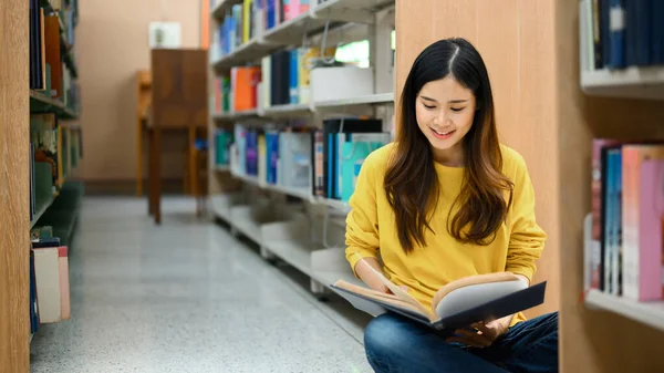 本の棚の近くの床に座っている若い女性に集中し テキストの本を読んで 研究を行い 試験を準備 — ストック写真