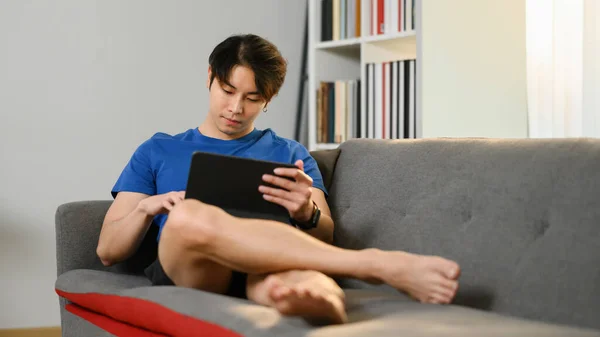 コンドームをつけません若いですアジア人男性でカジュアル着用でソファに座って インターネット上でデジタルタブレット — ストック写真