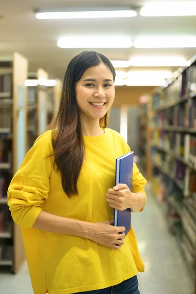 Portrait of university student woman holding textbook standing in library, looking at camera. Knowledge and education concept.