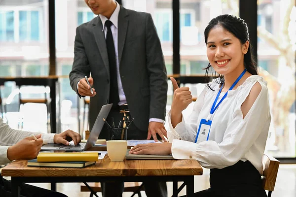 배경에 노트북 과그녀의 동료들을 일하는 미소짓는 아시아 엄지손가락을 — 스톡 사진