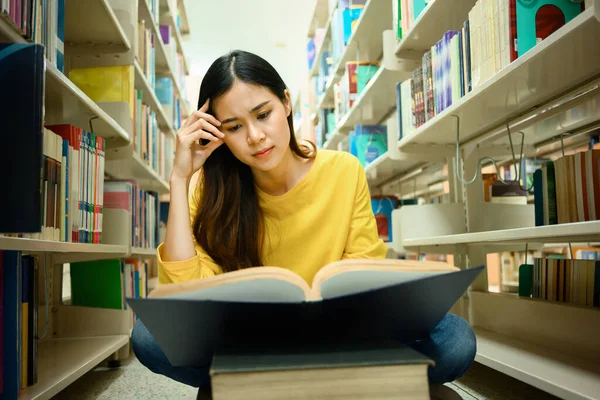 Confused female student sitting among bookshelves in library reading books for studying and research.