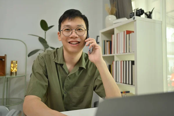 Mosolygó Ázsiai Férfi Szabadúszó Szemüvegben Beszél Mobiltelefonnal Használja Laptop Íróasztal — Stock Fotó