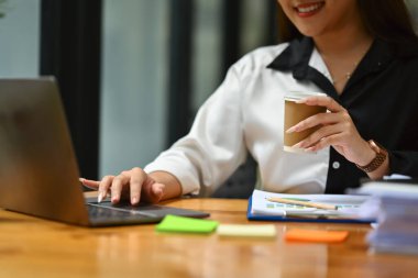 Elinde bir fincan kahve tutan ve bilgisayarını ahşap ofis masasında kullanan hoş, genç bir bayan çalışan..