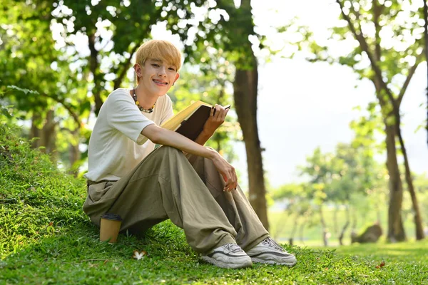 stock image Full length of cheerful young gay man reading book under tree on green grass at sunny beautiful day.