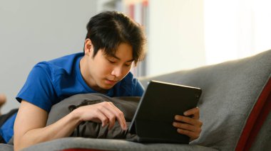 Kaygısız genç Asyalı adam kanepeye uzanıp internete veya internete dijital tabletle bakıyor..