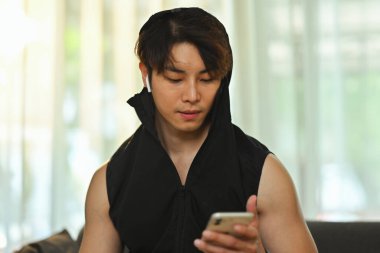 Milenyum Asyalı çekici bir adam sosyal medyada akıllı telefon iletişimini evde koltukta otururken kullanıyor..