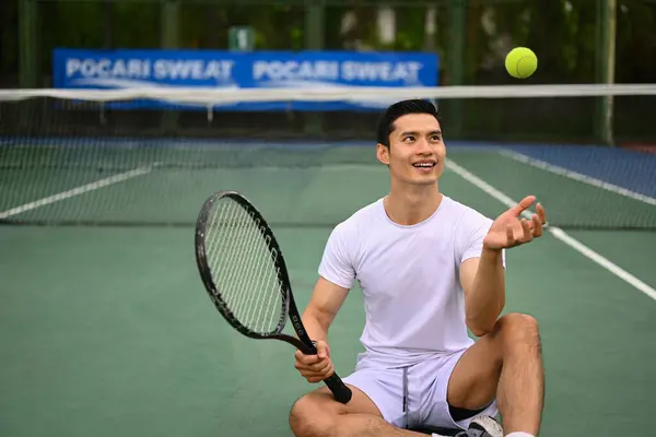 stock image Happy sportive asian man with racket throwing tennis ball in air, resting on tennis court. Exercise, sports and wellness concept.