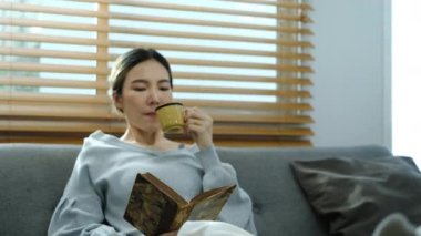 Hoş Asyalı kadın sabah kahvesini içiyor ve evdeki rahat koltukta kitap okuyor. Yavaş çekim.