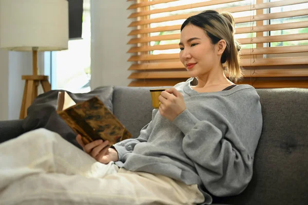 美丽的亚洲女子在客厅里喝着热茶 躺在沙发上看书 休闲和生活方式概念 — 图库照片