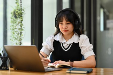 Konsantre Asyalı kız öğrenci kulaklık takıyor, dil öğreniyor, dizüstü bilgisayardan internetten webinar izliyor..