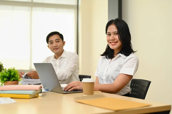 Confident young female secretary, office manager sitting in modern office with colleague and smiling to camera.