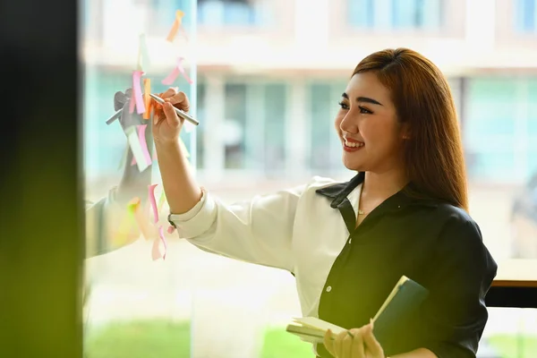 Smiling businesswoman standing near glass wall with colorful sticky papers, working on corporate project strategy in office.
