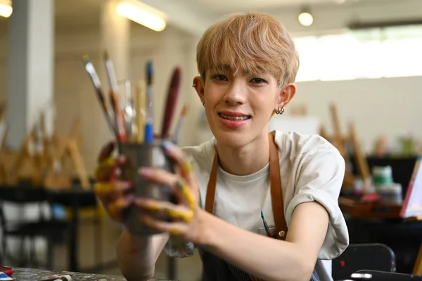 stock image Pretty young gay man dressed in apron holding metal bucket with different brushes of the artist, selective focus.