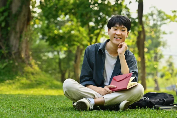 Uśmiechnięty Azjatycki Student Siedzący Trawie Parku Miejskim Czytający Książkę Styl — Zdjęcie stockowe