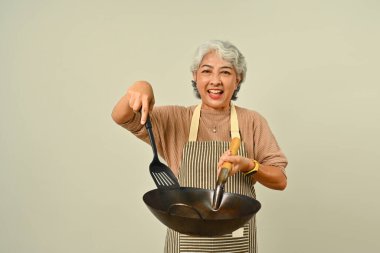 Neşeli 60 'lı yaşların beyaz saçlı kadın şefi önlük giyiyor elinde spatula ve kızarmış tava mavi arka planda izole edilmiş.. 