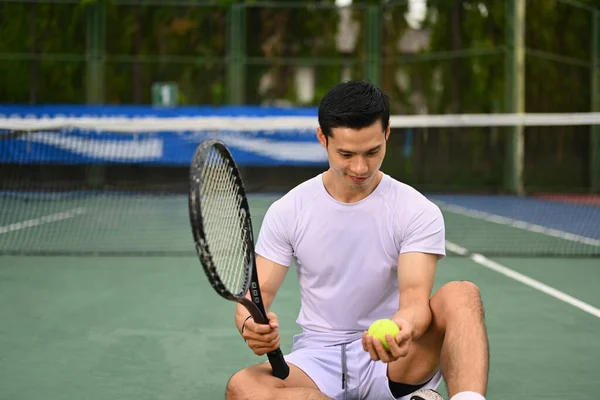 stock image Handsome Asian male tennis player in white sport clothes holding ball racket sitting on court.