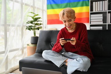 Oturma odasında duvarında gökkuşağı bayrağıyla oturmuş cep telefonuyla oyun oynayan neşeli gay adam..