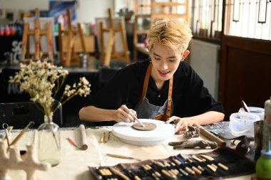 Odaklanmış genç adam çömlek çarkında kil tabağı yontuyor, sanat sınıfında el yapımı seramik yapıyor..