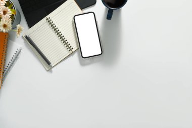 Boş ekranlı akıllı telefon, defter, kahve fincanı ve dizüstü bilgisayar beyaz masada. Üst görünüm, reklam metni için boş ekran.