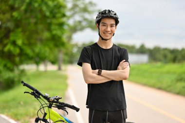 Bisiklet kıyafetleri ve kaskla bisiklet önünde kollarını kavuşturmuş gülümseyen bir adamın portresi..