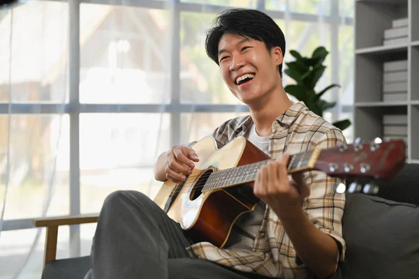 stock image Joyful young asian man playing acoustic guitar on couch, spending leisure time in cozy living room.