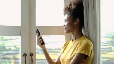 Relaxed young woman with Afro hair using smartphone and looking through window at modern cafe.