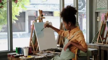 Casual young woman with Afro haired wearing apron painting picture, creating new artwork on canvas in art studio.