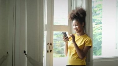 Happy young woman with curly haired using smartphone in bright living room. Communication, technology and lifestyle concept.