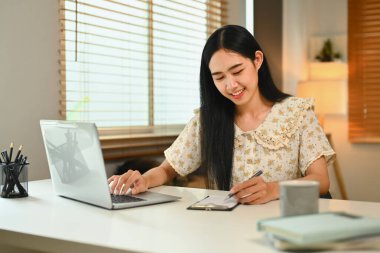Pretty young Asian woman using laptop and making notes, browsing internet, remote work and stying online.