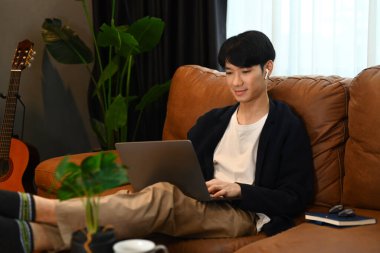 Kablosuz kulaklıklı, internette sörf yapan, dizüstü bilgisayarda çalışan rahat genç bir adam..