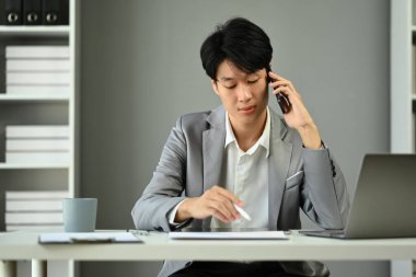 Asyalı erkek finansal danışmanın görüntüsü cep telefonu görüşmesi ve kağıt üzerinde bilgi okuma yoluyla müşteriye uzaktan danışma.