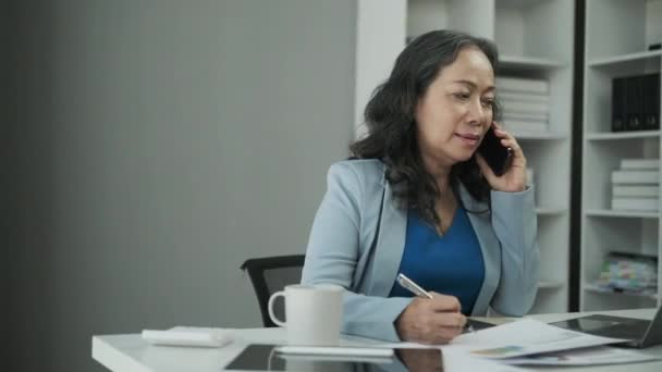 stock video Beautiful mature businesswoman having pleased phone conversation and taking notes on paper.