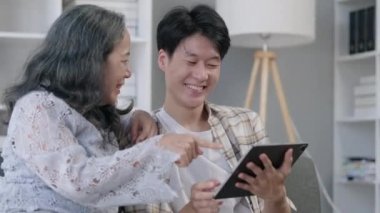 Kanepede oturup dijital tablet kullanan neşeli Asyalı genç adam ve büyükannenin yavaş çekimleri..