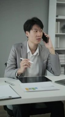 Asyalı erkek mali danışman cep telefonuyla ve kağıt üzerinde bilgi okuyarak müşteriye uzaktan danışmanlık yapıyor.